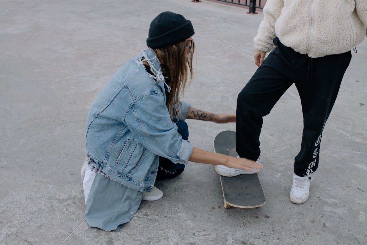 Two women interact with a skateboard on pavement, dressed in casual urban attire.