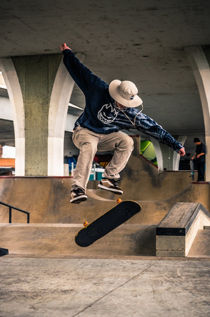 A skateboarder performs a high-energy trick in an urban skate park under a bridge.